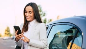 Car Insurance Banner - Businesswoman looking at her phone while standing outside her car.