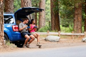 Car Insurance Banner - Two backpackers eating while sitting in the boot of their car in a forest