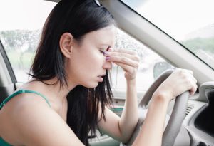 Car Insurance Banner - Woman pinches her nose in frustration while sitting at the wheel of her car.